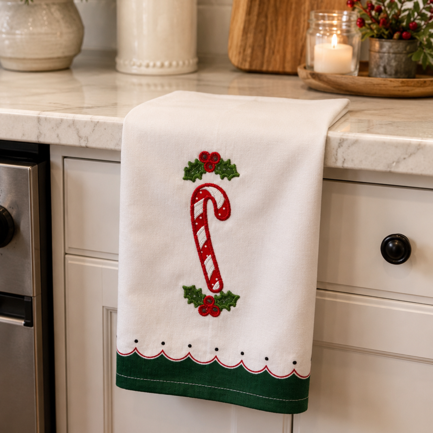 A white kitchen towel with a red and green embroidered candy cane and holly design, hanging over a white kitchen counter with kitchen utensils and plants in the background.