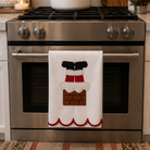 A white kitchen towel hanging from the handle of a stainless steel freestanding range, featuring an applique design of Santa's boots in a chimney stack. The background shows a white subway tile backsplash, a white pot on the stovetop, and wooden kitchen utensils.