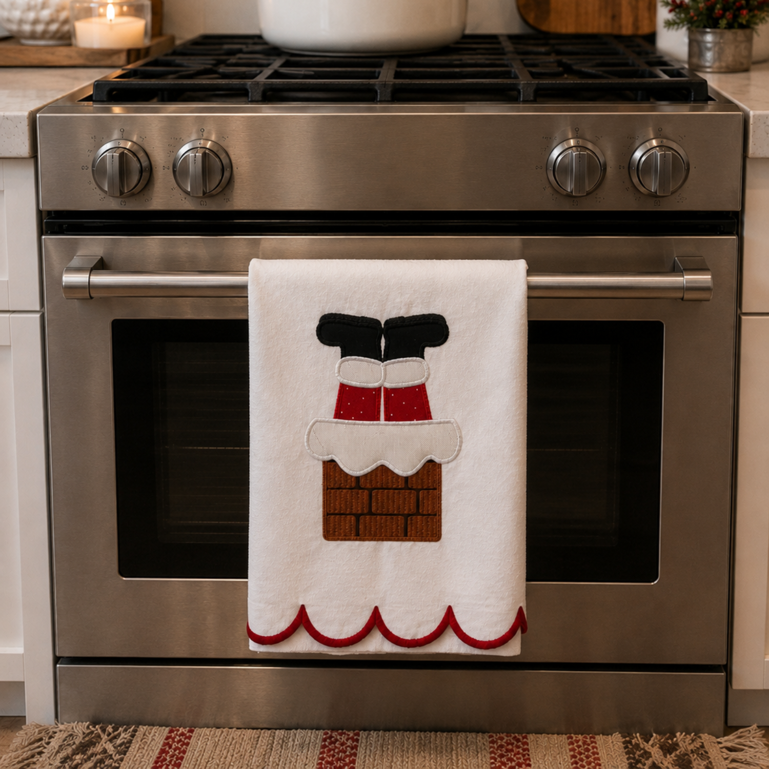 A white kitchen towel hanging from the handle of a stainless steel freestanding range, featuring an applique design of Santa's boots in a chimney stack. The background shows a white subway tile backsplash, a white pot on the stovetop, and wooden kitchen utensils.