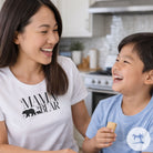 Woman wearing a 'Mama Bear' shirt laughing with a child in a kitchen.