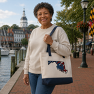 Woman holding a tote bag with a Maryland design by a waterfront with buildings and flags in the background.