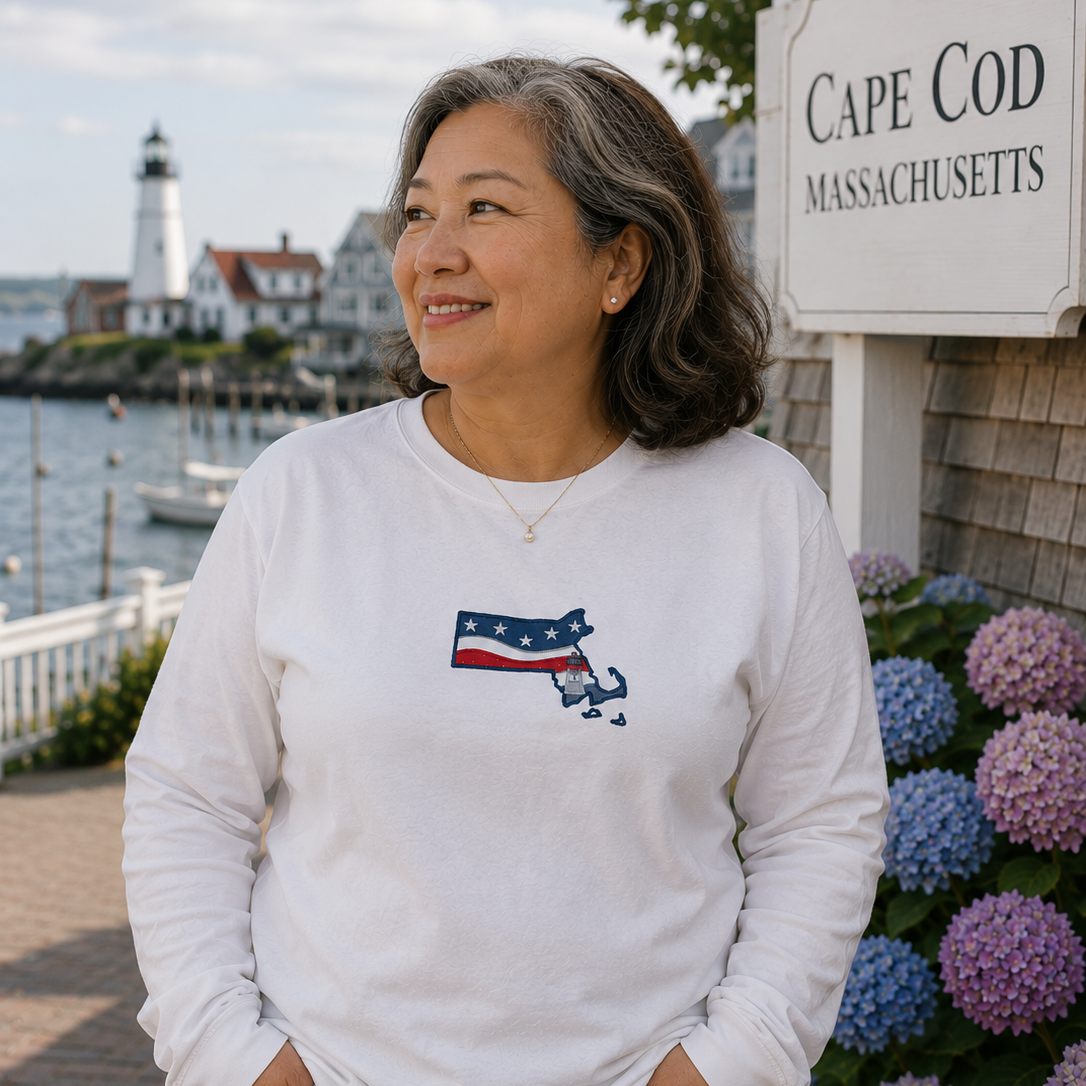 Woman wearing a white sweatshirt with a Cape Cod design, standing in front of a sign and water view.