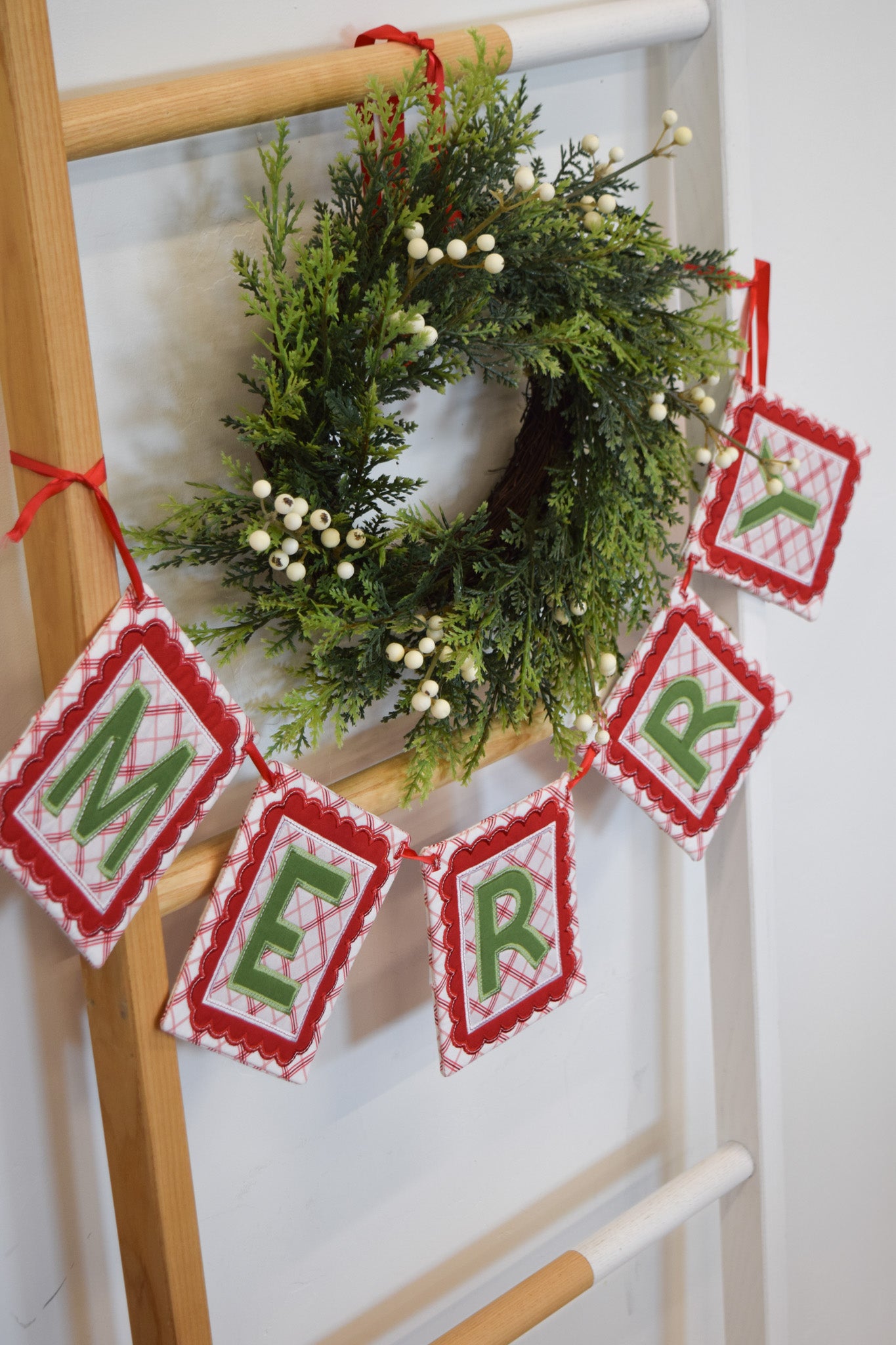 Christmas wreath with 'MERRY' banner on a wooden ladder against a white wall