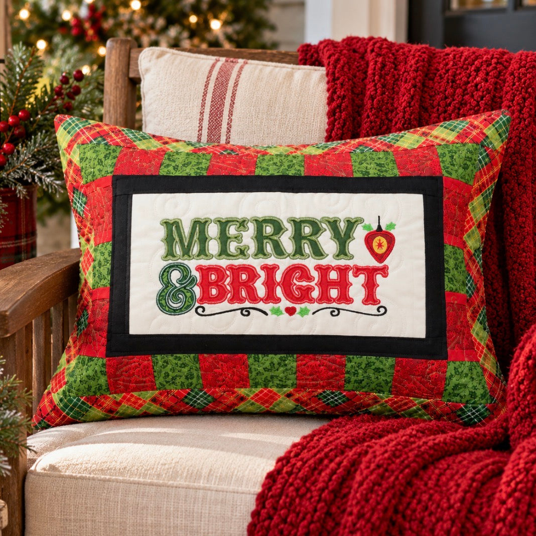 A square Christmas throw pillow with a red and green patchwork border and a central white panel reading “MERRY & BRIGHT” in green and red lettering, styled on a beige armchair with a red knit blanket and holiday decor in the background.