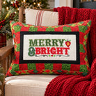 A square Christmas throw pillow with a red and green patchwork border and a central white panel reading “MERRY & BRIGHT” in green and red lettering, styled on a beige armchair with a red knit blanket and holiday decor in the background.