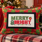 A square Christmas throw pillow with a red and green plaid border and a white center panel reading 'MERRY & BRIGHT' in green and red lettering, styled on a wooden bench with a red knit blanket and holiday decor in the background.