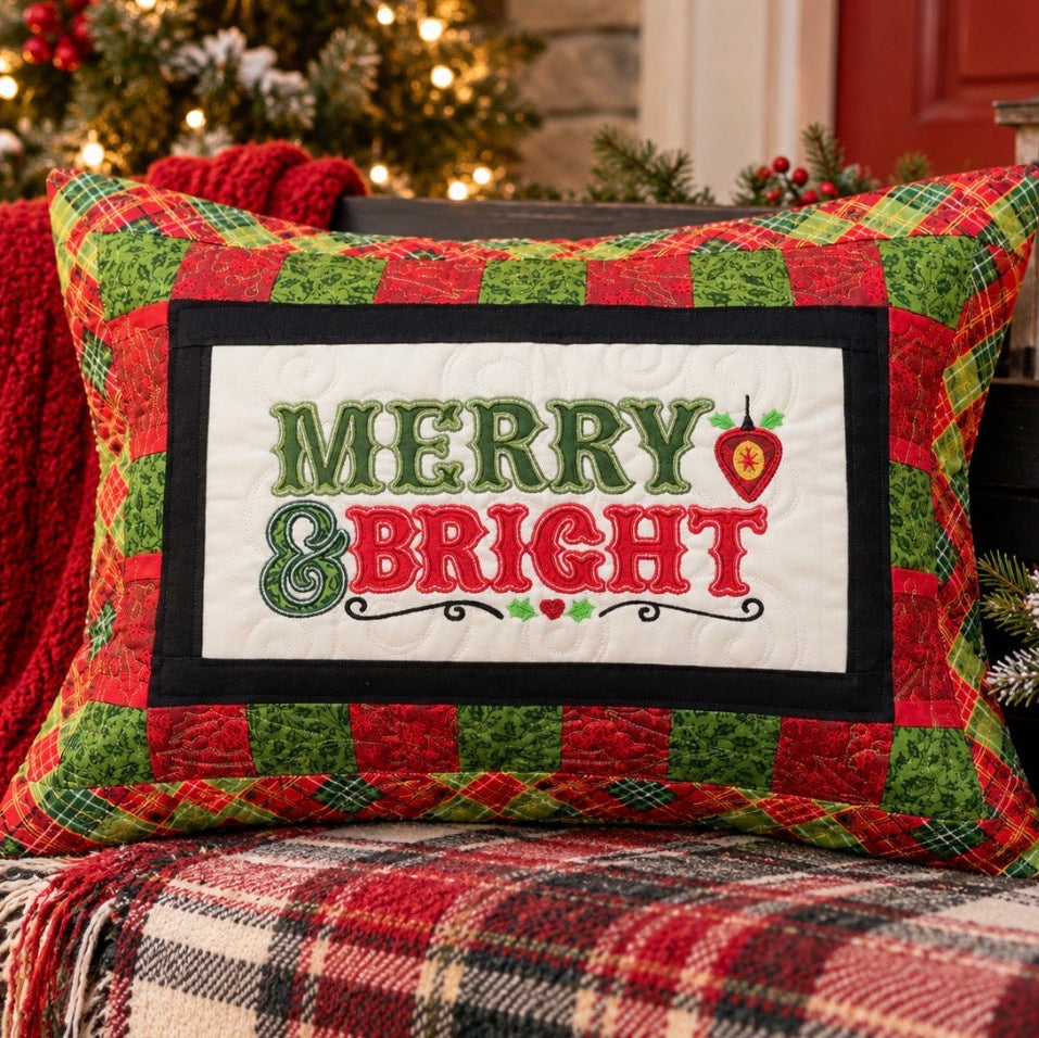 A square Christmas throw pillow with a red and green plaid border and a white center panel reading 'MERRY & BRIGHT' in green and red lettering, styled on a wooden bench with a red knit blanket and holiday decor in the background.