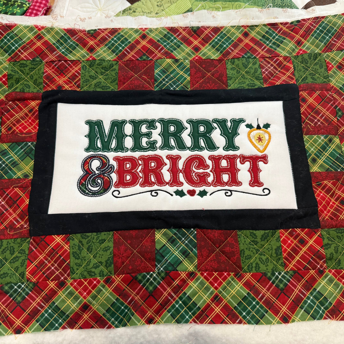 A handmade quilted Christmas table runner featuring a central white patch with 'MERRY & BRIGHT' embroidered in green and red, surrounded by a black border and a red and green plaid patchwork background.