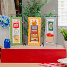 Three decorative kitchen towels displayed on a black wire stand on a red pedestal. The towels feature colorful autumn-themed illustrations including a jar of jam, a pumpkin, and a basket of red apples.