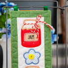 A close-up of a handmade quilted fabric wall hanging or decorative panel clipped to a metal rack. The panel features a green border, a white center, a red embroidered jar labeled "JAM" with a red-and-white striped ribbon, and a blue flower with a yellow center.