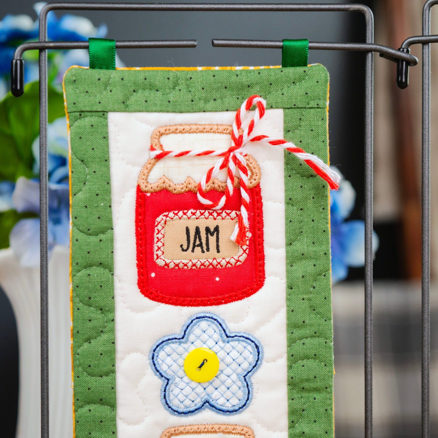 A close-up of a handmade quilted fabric wall hanging or decorative panel clipped to a metal rack. The panel features a green border, a white center, a red embroidered jar labeled "JAM" with a red-and-white striped ribbon, and a blue flower with a yellow center.