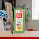A vertical quilted fabric banner with a honey-themed design, displayed on a black metal stand on a tabletop. The banner features a green border, a white center panel with a red honey jar, a blue flower, and a yellow honey pot.