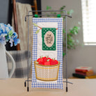 A kitchen towel with a blue and white gingham pattern, featuring a green banner that reads 'THE APPLE OF MY CARE' and an embroidered basket of red strawberries, hanging on a black metal stand on a tabletop.