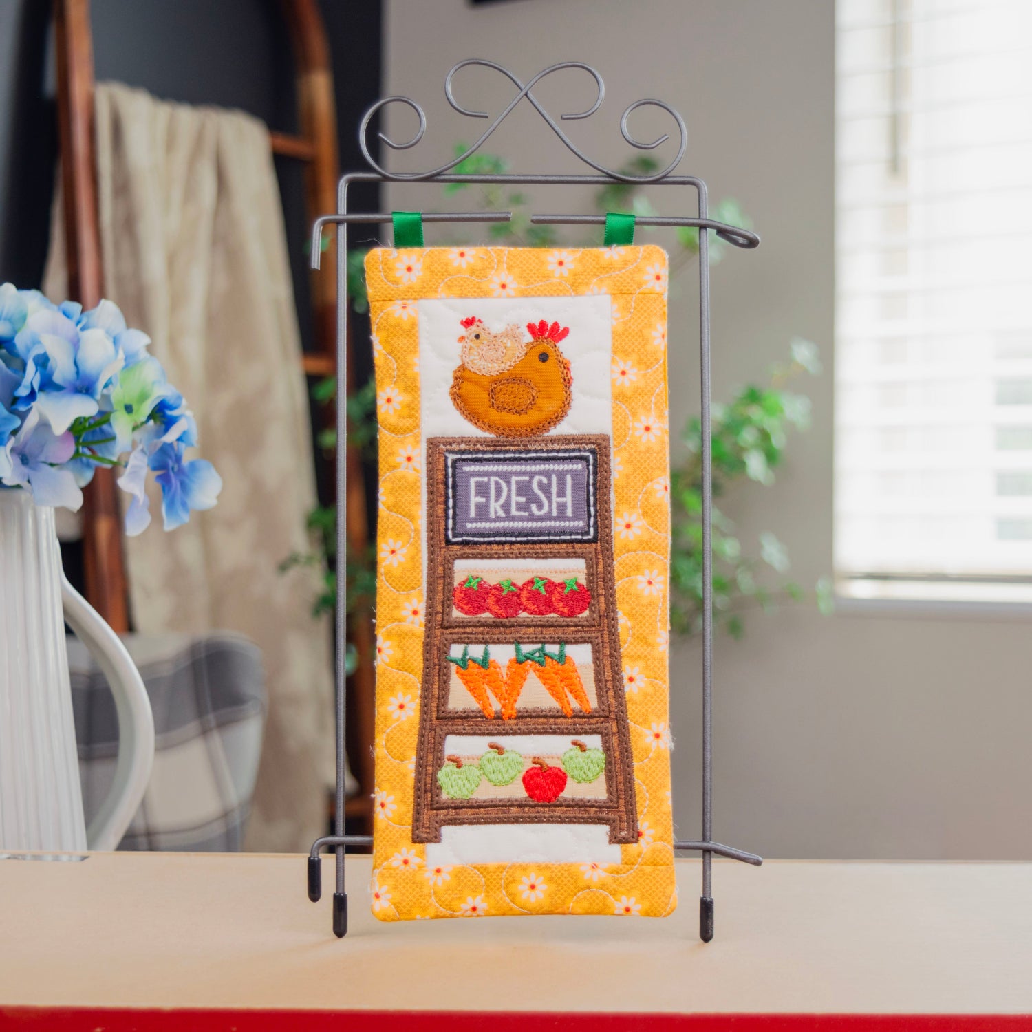 A decorative kitchen towel with a colorful embroidered chicken and produce display design, hanging on a black metal stand on a tabletop.