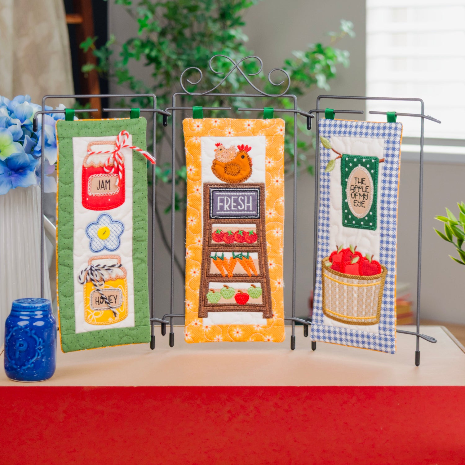 Three decorative kitchen-themed fabric pennant banners displayed on a black metal stand, set on a red box in a styled indoor scene with flowers, plants, and blue mason jars.