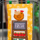 A close-up of a handmade quilted fabric hanging tag or pouch with a chicken and a chalkboard sign that reads 'FRESH', displayed on a metal rack.