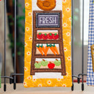 Three colorful quilted fabric garden stakes displayed on black metal stands. The center stake features a yellow floral border and an embroidered vegetable stand with red strawberries, orange carrots, green pears, and a brown sign reading 'FRESH'.