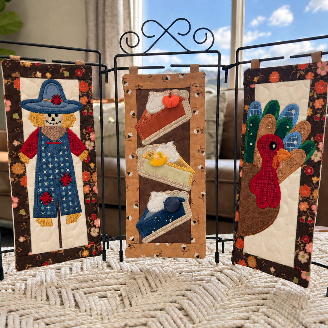 Three small quilted fabric wall panels displayed on a black metal stand in a home setting. The left panel shows a scarecrow, the center panel shows slices of pie, and the right panel shows a turkey.