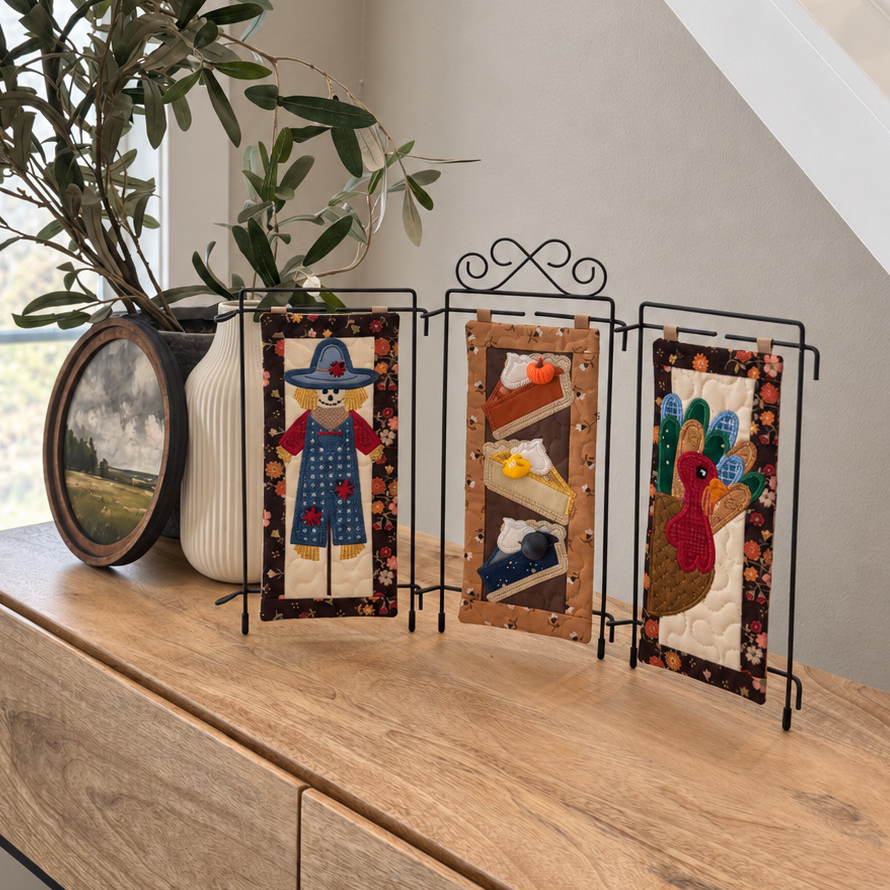Three small decorative quilted fabric wall art panels displayed on a black metal stand on a wooden console table in a home entryway, with a plant and staircase visible in the background.
