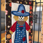A close-up of a handmade quilted fabric wall hanging featuring a stylized scarecrow. The scarecrow has a blue hat with a red flower, a white skull face, red sleeves, and blue overalls with white dots.