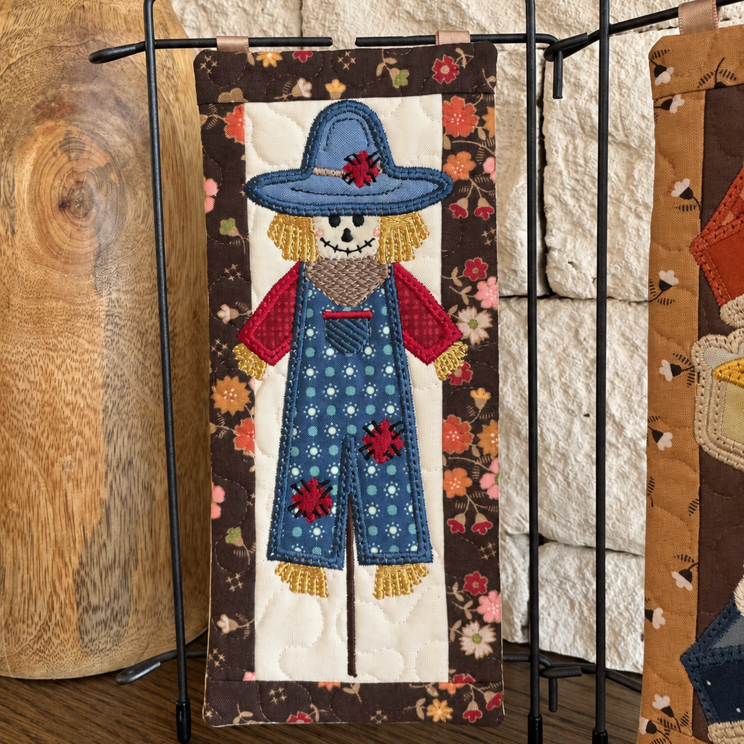A handmade quilted fabric banner featuring a stylized scarecrow applique, displayed on a black metal stand in front of a rustic stone wall and wood log.
