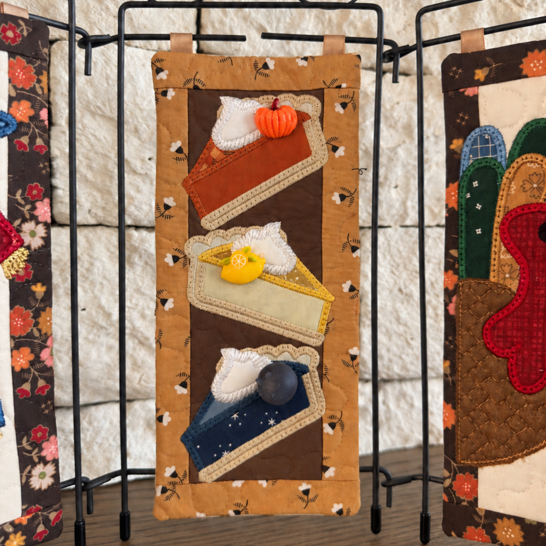 A handmade quilted fabric wall hanging or banner displayed on a black metal stand. The central panel features three stylized slices of pie with whipped cream and small decorative buttons, set against a tan and brown patterned background.