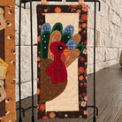 A handmade quilted fabric wall hanging featuring a turkey design, displayed on a black metal stand in front of a textured stone wall and wooden floor.