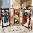 Three vertical fabric wall art panels displayed on a black metal stand in a home setting. The left panel shows a scarecrow in a blue outfit, the center panel shows stacked slices of pie, and the right panel shows a turkey with a red head and blue patterned wings.