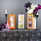 Three vertical acrylic wall art panels with Halloween-themed designs (ghosts, a witch's hat, potion bottles) displayed on a black and white spiderweb-patterned tablecloth next to a purple glass bottle and artificial flowers against a dark gray wall.