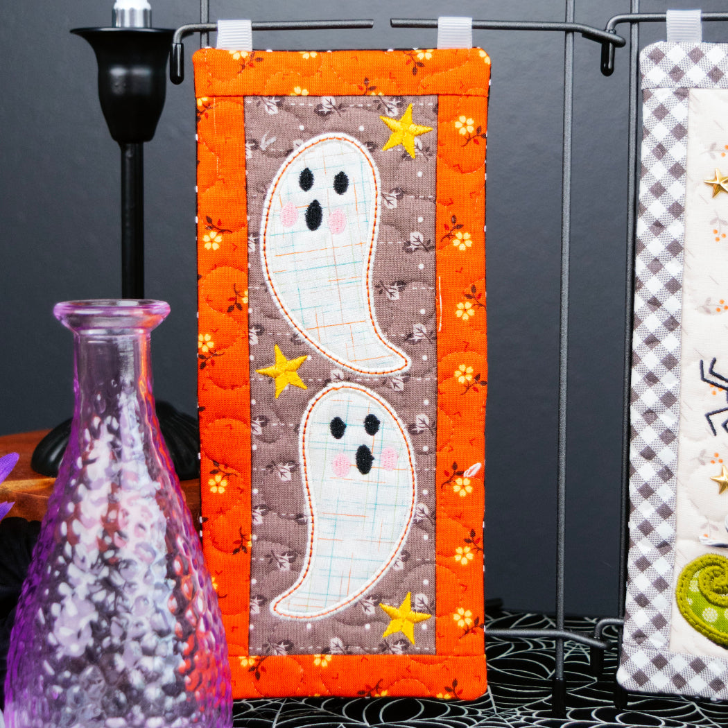 A Halloween-themed quilted fabric wall hanging with two white ghosts on a brown background, framed by an orange border with small yellow stars, displayed on a black stand against a dark backdrop.