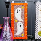 A Halloween-themed quilted fabric wall hanging with two white ghosts on a brown background, framed by an orange border with small yellow stars, displayed on a black stand against a dark backdrop.