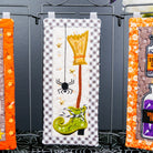 A Halloween-themed fabric tea towel or hanging cloth with a gingham border and embroidered witch motifs including a green boot, orange lantern, spider, and stars, displayed on a black metal stand between two similar orange-bordered cloths.