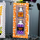 A Halloween-themed decorative sign featuring two glass potion bottles labeled 'Spider Legs' and 'Vampire Teeth' filled with purple liquid, set against a patterned orange and white background. The sign is displayed on a black metal stand on a table with a spiderweb pattern, with dark flowers in the background.