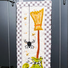 A Halloween-themed quilted wall hanging or tapestry displayed on a black metal rack. The central panel features a white and gray gingham background with a stitched yellow witch's hat, a black spider, gold stars, and a green boot at the bottom.