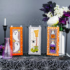 Three Halloween-themed fabric wall banners displayed on a black wire stand on a table. The banners feature orange borders and whimsical designs: two white ghosts on the left, a witch's hat and green boot on the center, and potion bottles on the right.