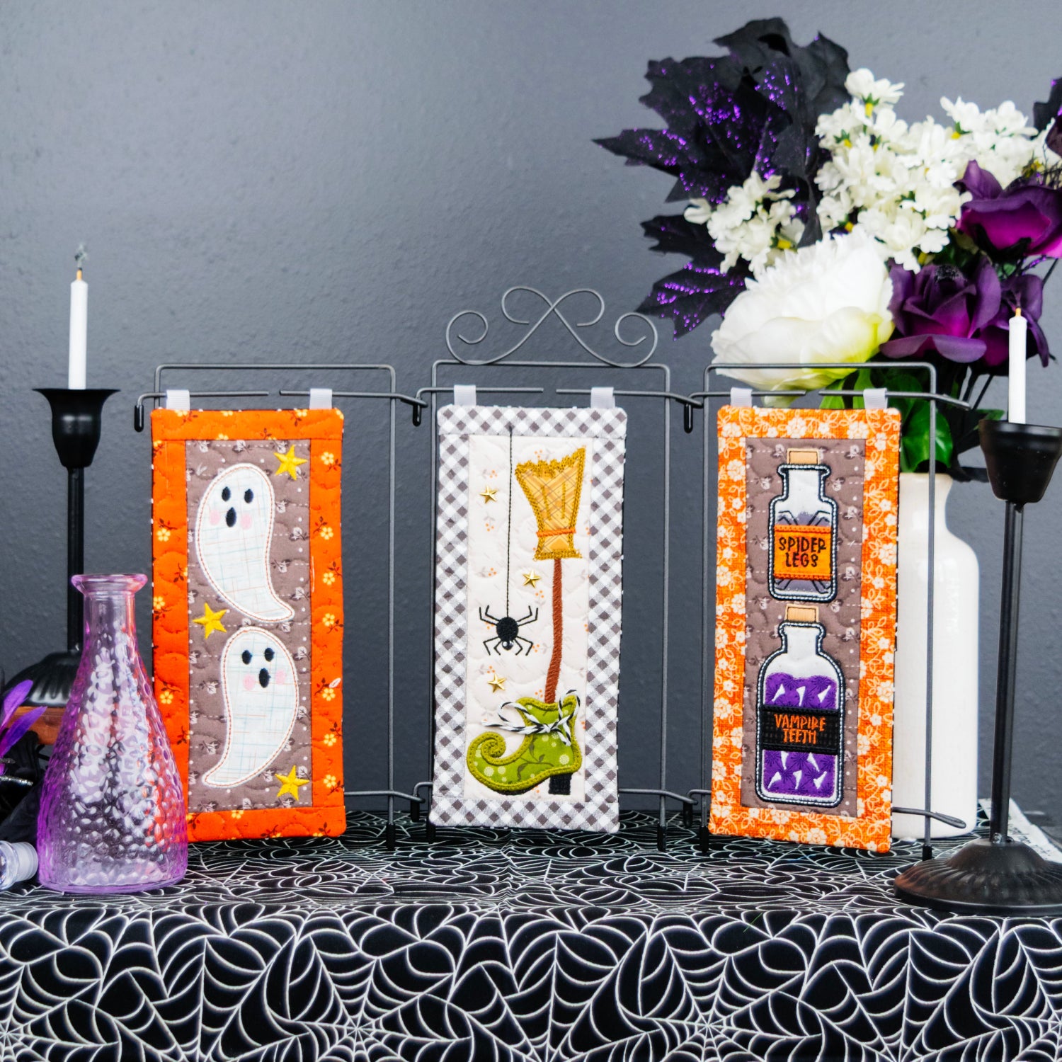 Three Halloween-themed fabric wall banners displayed on a black wire stand on a table. The banners feature orange borders and whimsical designs: two white ghosts on the left, a witch's hat and green boot on the center, and potion bottles on the right.