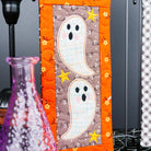 Two Halloween-themed fabric wall hangings displayed on a dark wall. The left hanging features two stitched ghosts on an orange and brown patterned background, and the right hanging shows a spider, a green boot, and a yellow lantern on a white checkered background.