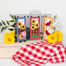 Decorative kitchen towels with sunflower and watermelon designs on a stand with a red and white checkered tablecloth.