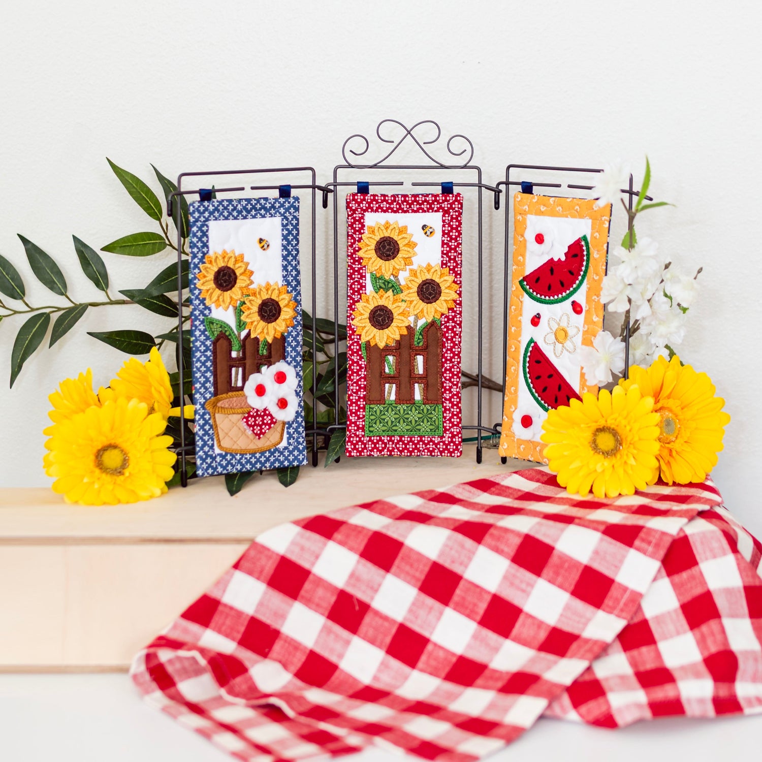 Decorative kitchen towels with sunflower and watermelon designs on a stand with a red and white checkered tablecloth.