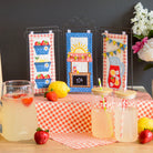 Table setting with lemonade, fruits, and decorative items on a checkered tablecloth.