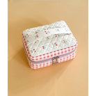 A small quilted fabric zip case with a white top panel printed with tiny cherry motifs, a pink-and-white gingham border, and a silver zipper closure, shown on a warm beige background.
