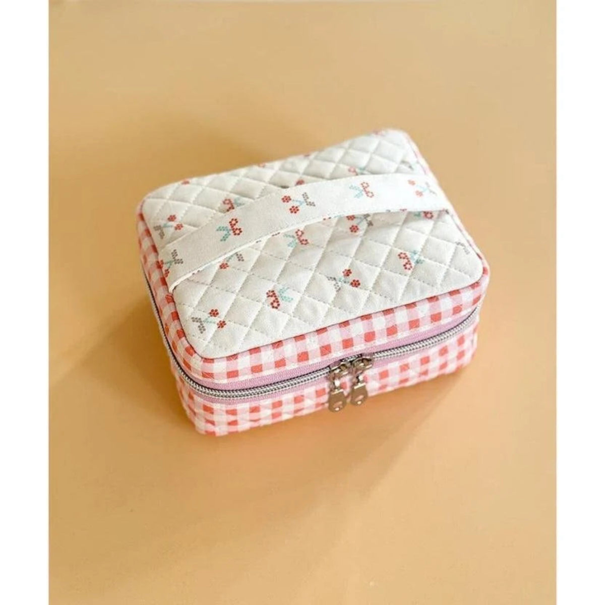 A small quilted fabric zip case with a white top panel printed with tiny cherry motifs, a pink-and-white gingham border, and a silver zipper closure, shown on a warm beige background.