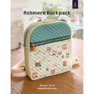 An Ashmere backpack shown on a wooden table in a softly blurred indoor setting. The backpack has a quilted design with a teal polka-dot top panel, a patterned lower section with small house motifs, and a light beige strap.