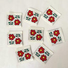 Seven small square white tiles or stickers arranged on a light beige background, each printed with a red flower motif and green leaves. A small stylized logo appears in the bottom right corner.