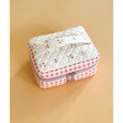 A small quilted zippered pouch with a cherry print on the top and a red-and-white gingham border, shown on a warm beige background.