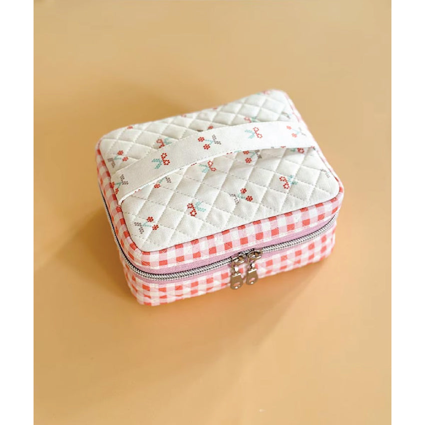 A small quilted zippered pouch with a cherry print on the top and a red-and-white gingham border, shown on a warm beige background.