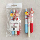 Three cute heat erasable pens with cartoon character charms are shown, with one pen in its clear plastic packaging and the other two standing beside it on a light beige fabric background.