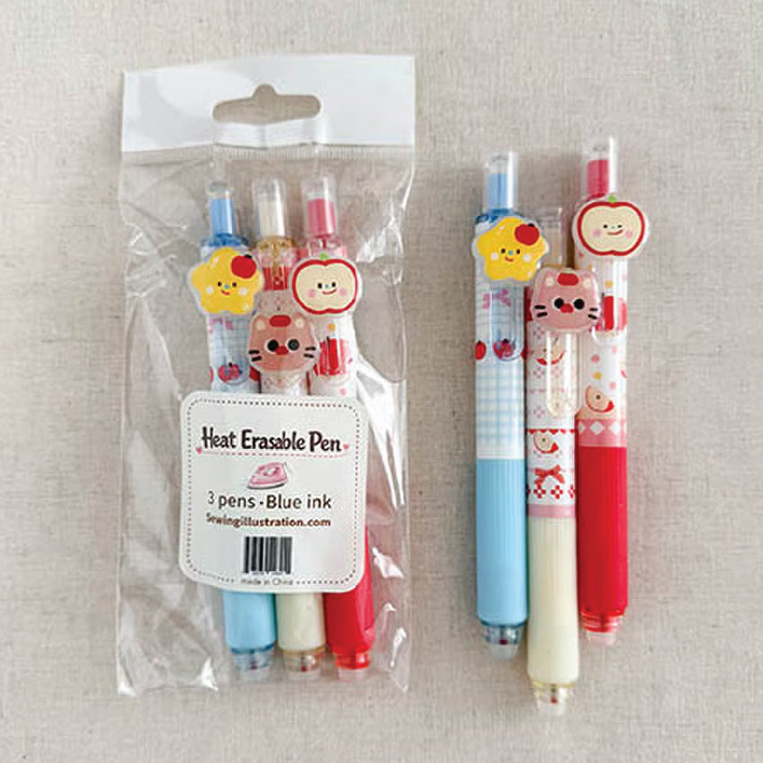 Three cute heat erasable pens with cartoon character charms are shown, with one pen in its clear plastic packaging and the other two standing beside it on a light beige fabric background.