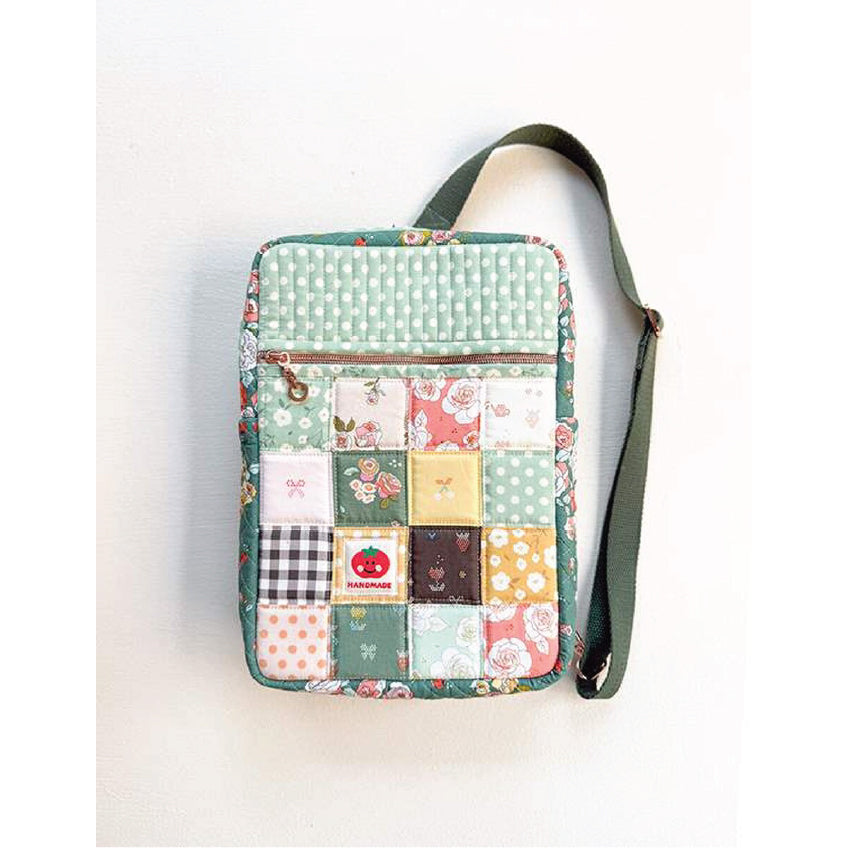 A square quilted crossbody bag with a patchwork design featuring various floral, polka dot, and solid fabric squares in green, pink, yellow, and white. The bag has a top zipper, a green fabric strap, and a small decorative zipper pull with a cartoon character.
