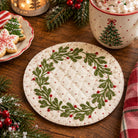 A round quilted coaster with a green holly wreath and red berries on a cream background, styled on a wooden table with holiday decorations, a mug, and cookies.